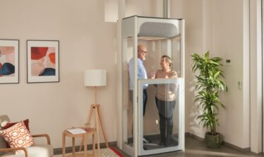 Starší pár stojaci v domácom výťahu Stannah Uplifts vo svetlej obývačke, ktorý sa vyznačuje kompaktným dizajnom prechádzajúcim cez podlahu s priehľadnými panelmi.