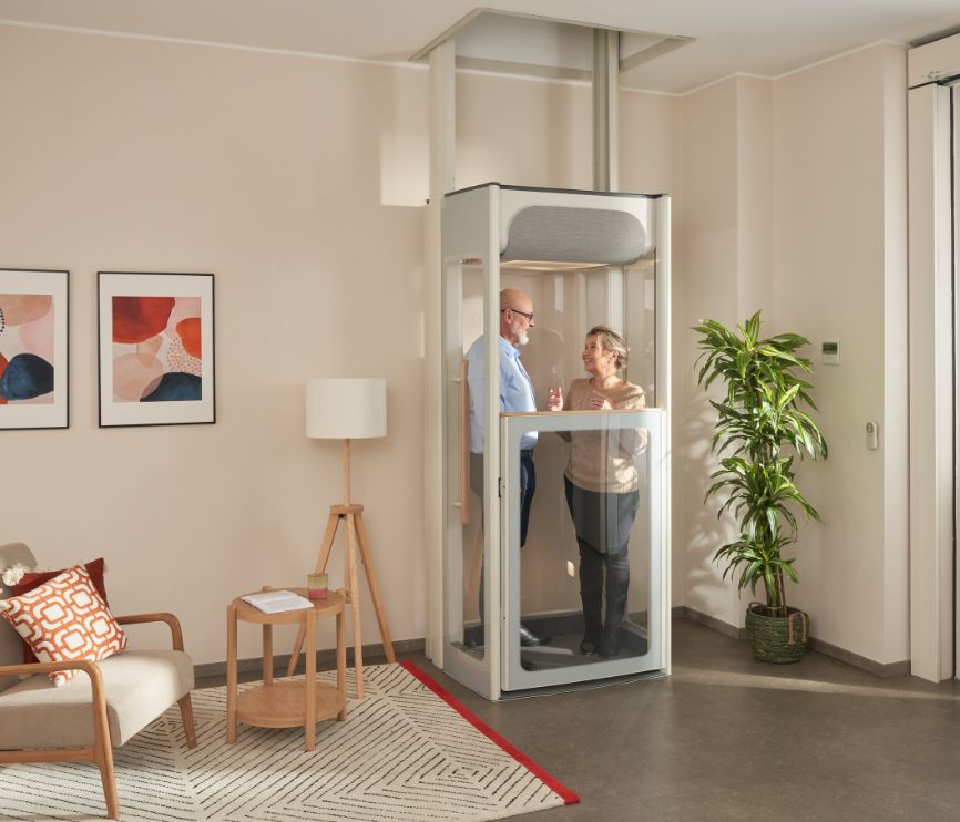 Starší pár stojaci v domácom výťahu Stannah Uplifts vo svetlej obývačke, ktorý sa vyznačuje kompaktným dizajnom prechádzajúcim cez podlahu s priehľadnými panelmi.